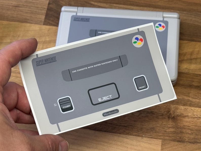 May include: A grey and white Super Nintendo console sticker is held up, with another console visible in the background. The sticker shows the console's design, including the "EJECT" button and the Nintendo logo.