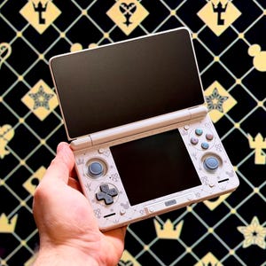 Op de afbeelding: Een witte Nintendo 3DS handheld gameconsole met een zwart scherm, open en in een hand gehouden. De console heeft lichtblauwe knoppen en een decoratief patroon. De achtergrond is zwart met goudkleurige symbolen.