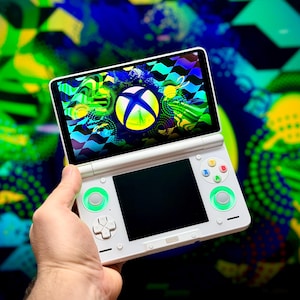 Puede incluir: Consola de juegos portátil blanca con una pantalla que muestra un gráfico colorido con un logotipo de Xbox. La consola tiene detalles en verde en los botones de control y una pantalla negra debajo de la pantalla principal.
