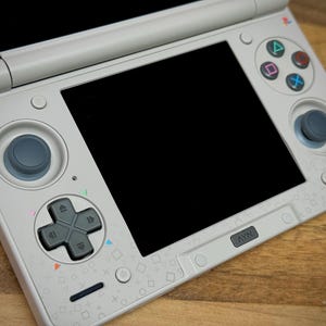 May include: A white Sony handheld gaming console with a black screen, directional pad, and action buttons. The console has a gray joystick and a NAV button. The surface is decorated with small, colorful geometric shapes.