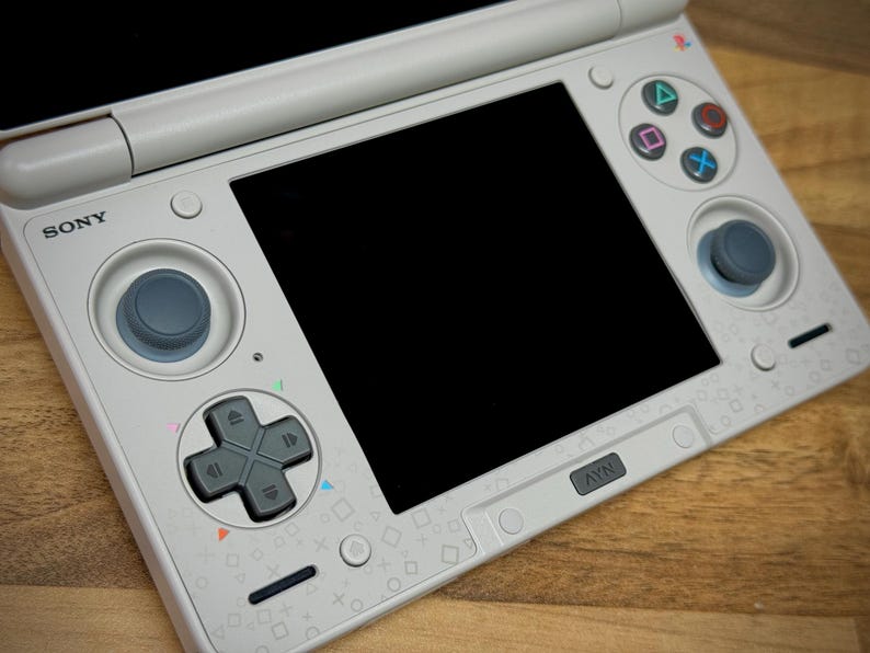 May include: A white Sony handheld gaming console with a black screen, directional pad, and action buttons. The console has a gray joystick and a NAV button. The surface is decorated with small, colorful geometric shapes.