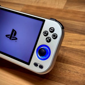 Puede incluir: Consola de juegos portátil plateada con una pantalla azul que muestra el logotipo de PlayStation. La consola tiene botones negros y un joystick iluminado en azul. El dispositivo está sobre una superficie de madera.