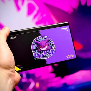 May include: Black Nintendo Game Boy Advance SP console with a purple and black design featuring a Gengar graphic and Japanese text. The console is held in a hand against a vibrant purple and pink background.