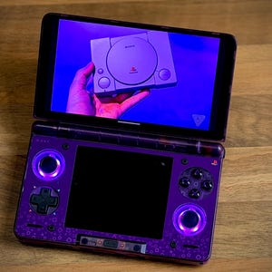 May include: A purple handheld gaming console with a screen displaying a PlayStation console. The console has a black screen, purple buttons, and glowing purple lights. The background is a wooden surface.