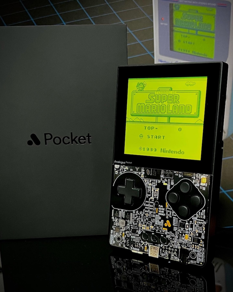 May include: A black handheld gaming device with a green screen displaying the game "Super Mario Land". The device has a cross-shaped button and four round buttons. The text "Analogue Pocket" and "1939 Nintendo" are visible on the screen.