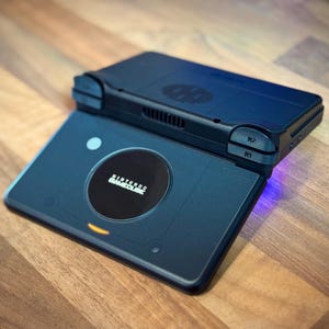 May include: Two dark blue handheld gaming consoles stacked on a wooden surface. The bottom console has a circular design with the text "NINTENDO GAMECUBE". The top console has buttons labeled "R1" and "R2".