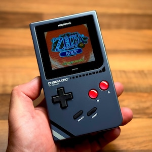 Può includere: Console di gioco portatile grigio scuro con schermo che mostra "The Legend of Zelda: Oracle of Ages". Ha un D-pad nero, pulsanti rossi e la scritta "Chromatic 1st Edition".