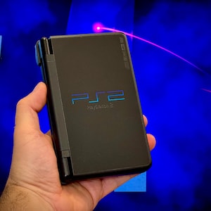 May include: A black PlayStation 2 handheld console, held in a hand, with the PS2 logo in blue and the text "PlayStation 2". The background is a blurry blue with a red laser beam.