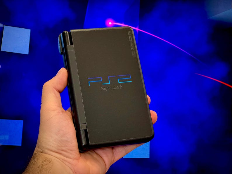 May include: A black PlayStation 2 handheld console, held in a hand, with the PS2 logo in blue and the text "PlayStation 2". The background is a blurry blue with a red laser beam.