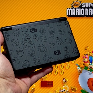 Puede incluir: Una consola de juegos portátil con una cubierta negra adornada con personajes e iconos de Mario. El fondo es naranja con el texto "New Super Mario Bros. 2".