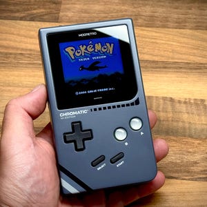May include: A gray handheld gaming device displaying the "Pokémon Silver Version" game. The device has a black screen, a directional pad, and action buttons. The text "CHROMATIC 1st EDITION" is visible on the device.