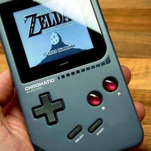 Könnte beinhalten: Graues Handheld-Spielgerät mit "The Legend of Zelda: Link's Awakening" auf dem Bildschirm. Es hat ein Steuerkreuz, rote Knöpfe und den Text "CHROMATIC 1st EDITION".