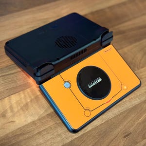 Puede incluir: Una consola portátil Nintendo GameCube naranja y negra. La consola tiene una parte superior negra y una inferior naranja con un círculo negro y las palabras "Nintendo GameCube". La consola está sobre una superficie de madera.