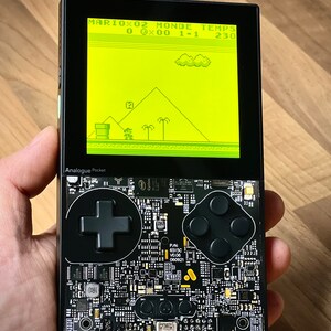 May include: A black handheld gaming device with a yellow screen displaying a video game. The screen shows the text "MARIOX02 MONDE TEMPS 0 0x00 1-1 230". The device has a black directional pad and four black buttons.