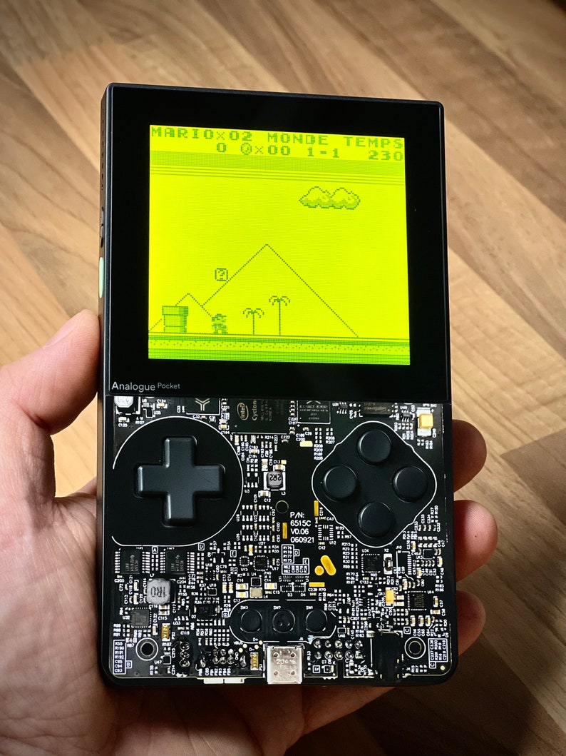 May include: A black handheld gaming device with a yellow screen displaying a video game. The screen shows the text "MARIOX02 MONDE TEMPS 0 0x00 1-1 230". The device has a black directional pad and four black buttons.