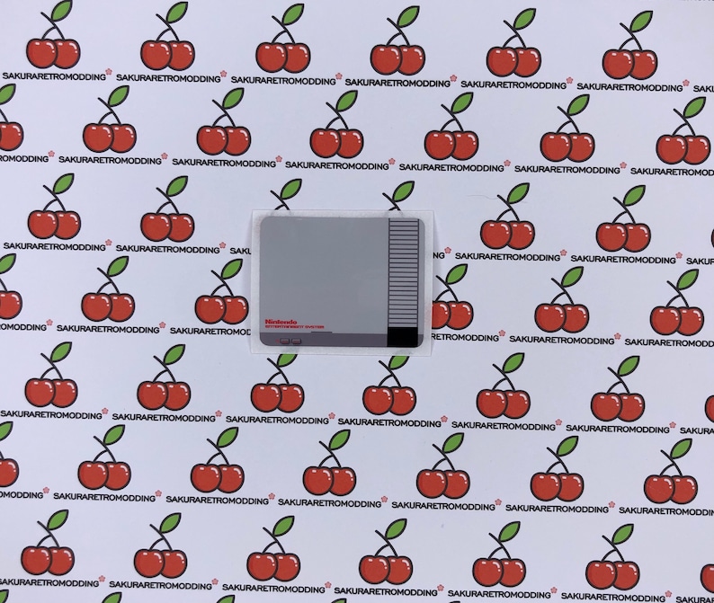 Puede incluir: Primer plano de un cartucho gris de Nintendo Entertainment System con una etiqueta blanca. La etiqueta tiene el texto "Nintendo Entertainment System" y una peque&ntilde;a imagen de un mando. El cartucho est&aacute; rodeado de un fondo blanco con un patr&oacute;n repetitivo de cerezas rojas.