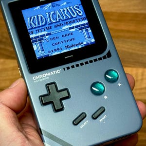 Könnte beinhalten: Handheld-Spielkonsole in metallischem Blau-Grau, die das Spiel "Kid Icarus" auf dem Bildschirm zeigt. Das Gerät hat ein Steuerkreuz, grüne Knöpfe und den Text "CHROMATIC 1st EDITION".