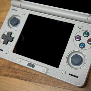 May include: A light gray handheld gaming console with a black screen and various buttons, including a directional pad, analog stick, and action buttons. The console has the Sony logo and a PlayStation symbol.