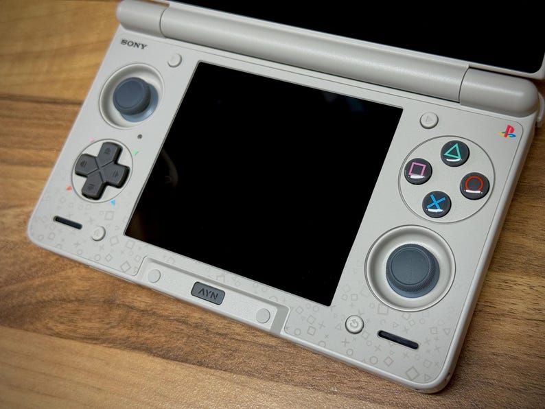 May include: A light gray handheld gaming console with a black screen and various buttons, including a directional pad, analog stick, and action buttons. The console has the Sony logo and a PlayStation symbol.