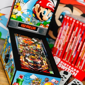 Op de afbeelding: Een miniatuur flipperkast met een zwart frame en een kleurrijk Mario-thema. De machine heeft een digitaal display dat de score "162050130" laat zien. De flipperkast is verlicht met een fel oranje licht.