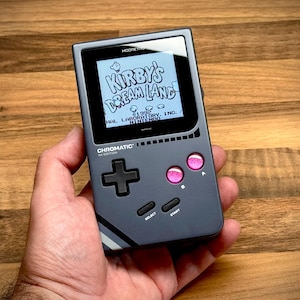 Könnte beinhalten: Ein graues Handheld-Spielgerät, das "Kirby's Dream Land" auf dem Bildschirm anzeigt. Das Gerät hat rosa Knöpfe, ein schwarzes Steuerkreuz und den Text "CHROMATIC" und "1st EDITION".