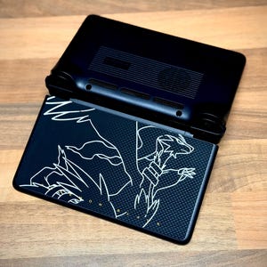 May include: Black handheld gaming console, open to reveal a screen with a white graphic design. The design features a dragon-like creature. The console is on a wooden surface.