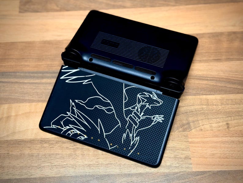 May include: Black handheld gaming console, open to reveal a screen with a white graphic design. The design features a dragon-like creature. The console is on a wooden surface.