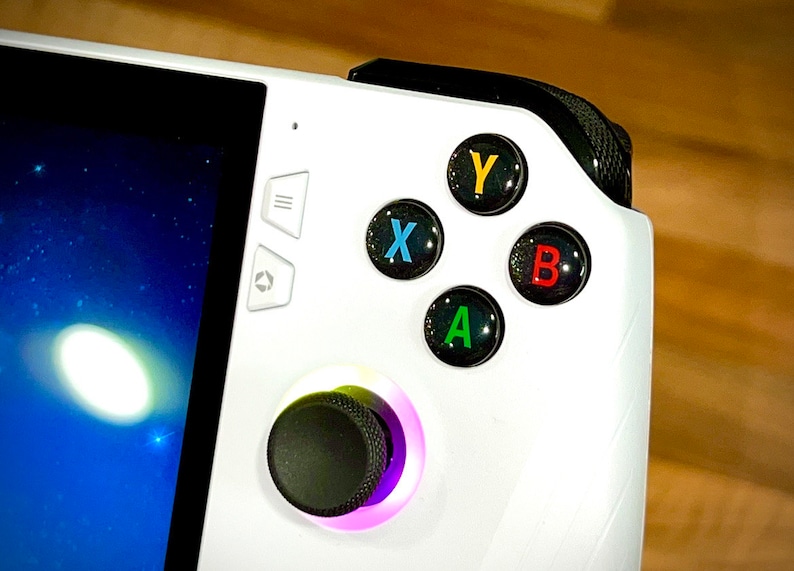 May include: A white handheld gaming device with a black trim. The device has a black and purple joystick, a black button with a white 'A' on it, a black button with a green 'B' on it, a black button with a blue 'X' on it, and a black button with a yellow 'Y' on it. The device has a screen with a blue background and white stars.