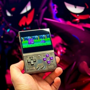 May include: A handheld retro gaming console with a purple and green pixelated game displayed on the screen. The console has a black and silver exterior with purple buttons. The console is held in a hand against a background of purple and red.