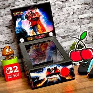 Peut inclure: Mini-flipper sur le thème de "Retour vers le futur", avec un graphisme coloré de l'affiche du film. Comprend une console Nintendo Switch, une figurine Goomba et une décoration en forme de cerise. Des manettes de jeu noires et oranges sont également présentes.