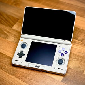 May include: A white handheld gaming console with a black screen and a hinged top. It features a directional pad, buttons, and a joystick. The console is open, displaying two screens, and rests on a wooden surface.