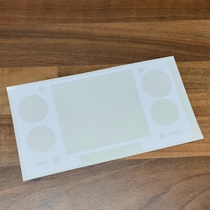 Pu&ograve; includere: Un foglio di adesivi bianco con uno schermo rettangolare verde chiaro e ritagli circolari per i pulsanti, progettato per un dispositivo di gioco portatile. L'adesivo &egrave; su una superficie di legno.