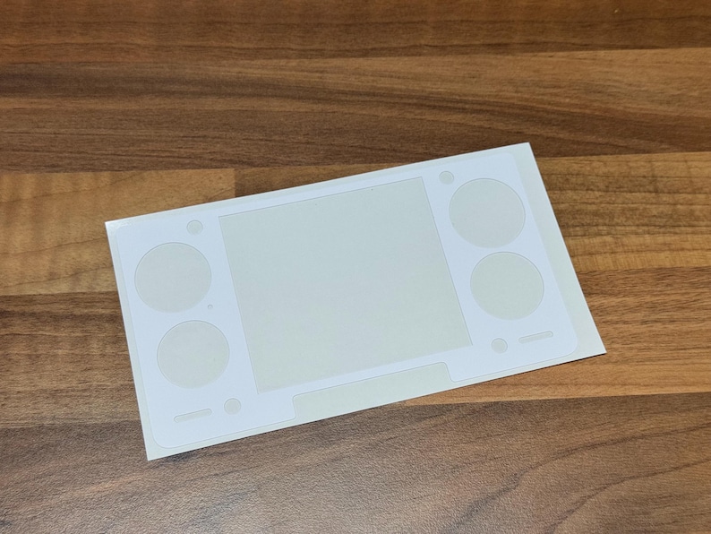 Pu&ograve; includere: Un foglio di adesivi bianco con uno schermo rettangolare verde chiaro e ritagli circolari per i pulsanti, progettato per un dispositivo di gioco portatile. L'adesivo &egrave; su una superficie di legno.