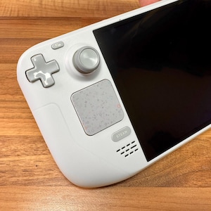 May include: A white handheld gaming device with a black screen, a gray directional pad, and a gray thumbstick. The device has a gray touchpad and the word "STEAM" printed on the bottom. The device is on a wooden surface.