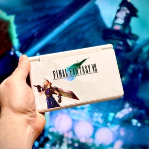 Könnte beinhalten: Weiße Nintendo DS Lite Handheld-Spielkonsole mit dem Final Fantasy VII-Logo und Charakterillustration. Die Konsole wird in einer Hand gehalten, mit einem verschwommenen Hintergrund einer Videospielszene.