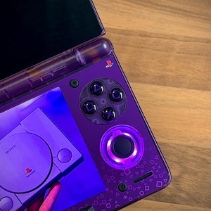 Peut inclure: Console de jeu portable violette personnalisée sur le thème de la PlayStation. La console présente une coque translucide violette, un pavé directionnel circulaire et la disposition classique des boutons PlayStation. Une console PlayStation 1 est affichée.