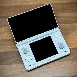May include: A white handheld gaming console with two screens, one open and one closed. The console has a directional pad, buttons, and a joystick on the bottom half. The top screen is open, displaying a black screen. The console is on a wooden surface.