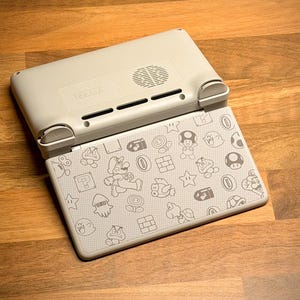 May include: A light gray handheld gaming console with a patterned design featuring characters and icons from the Super Mario Bros. series. The console is open, revealing the screen with the design. The device is on a wooden surface.