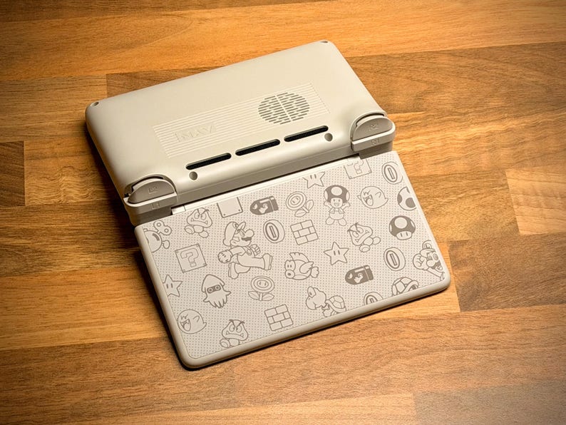 May include: A light gray handheld gaming console with a patterned design featuring characters and icons from the Super Mario Bros. series. The console is open, revealing the screen with the design. The device is on a wooden surface.
