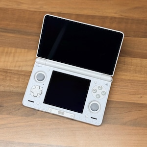 Könnte beinhalten: Eine weiße tragbare Spielkonsole mit zwei Bildschirmen, bestehend aus einem oberen Display und einem unteren Touchscreen. Das Gerät verfügt über ein Steuerkreuz, zwei Analogsticks und vier Aktionstasten auf der Vorderseite, Sony-Logo sichtbar.