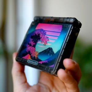 Peut inclure: Une console de jeu portable avec un écran affichant un motif de vague rétro rose, violet et bleu. L'appareil a un cadre noir et le logo Nintendo est visible.