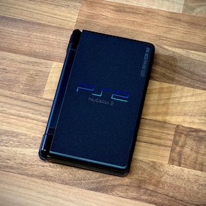 May include: A black PlayStation 2 console, featuring the PlayStation 2 logo in blue. The console is closed and resting on a wooden surface. The image highlights the sleek design of the gaming console.