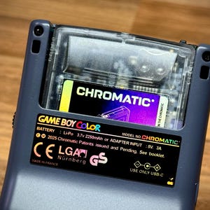 May include: Close-up of a Game Boy Color handheld console with a "CHROMATIC" cartridge inserted. The console is dark gray with a clear plastic cartridge slot. The cartridge has a black, yellow, and purple label. Text on the console includes "GAME BOY COLOR" and "CHROMATIC".