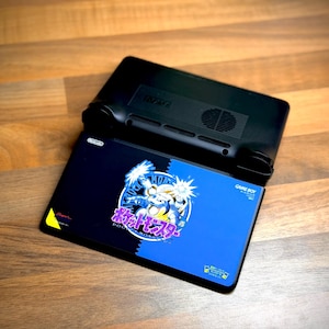 May include: Black handheld gaming console with a blue and black game cartridge featuring a cartoon character and Japanese text. The console is resting on a wooden surface.