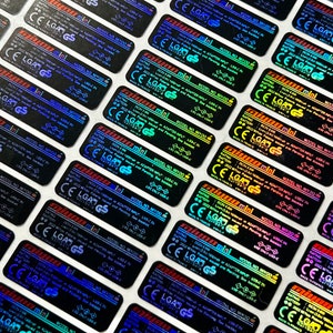 K&ouml;nnte beinhalten: Nahaufnahme von mehreren holografischen Aufklebern mit dem Text "mini" und "MODEL NO. MY700". Die Aufkleber sind schwarz mit einem regenbogenfarbenen, irisierenden Finish.