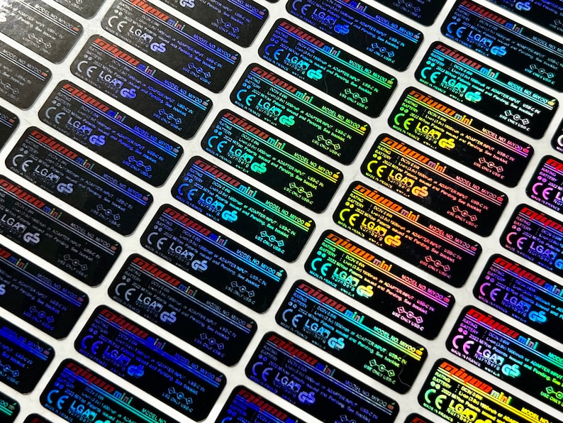 K&ouml;nnte beinhalten: Nahaufnahme von mehreren holografischen Aufklebern mit dem Text "mini" und "MODEL NO. MY700". Die Aufkleber sind schwarz mit einem regenbogenfarbenen, irisierenden Finish.