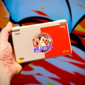 May include: A beige and red Nintendo DS Lite handheld game console featuring a "Monster" graphic with a dragon-like creature and Japanese text. The console has a "Game Boy" sticker and is held in a hand.