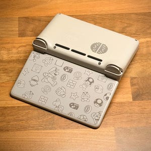 May include: A light gray handheld gaming console with a patterned cover featuring cartoon characters. The console is open, revealing the screen with a design of Mario and other game elements. The top part of the console is light gray with vents and a speaker.
