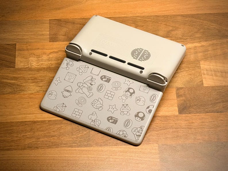 May include: A light gray handheld gaming console with a patterned cover featuring cartoon characters. The console is open, revealing the screen with a design of Mario and other game elements. The top part of the console is light gray with vents and a speaker.