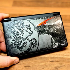 May include: A black handheld gaming console featuring a detailed graphic on the top cover. The design includes a circular tribal-style emblem on the left and a large, textured creature illustration on the right with a prominent red accent mark.